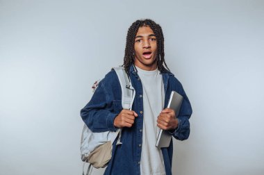 Genç bir öğrenci tek eli ve sırtında bir sırt çantasıyla dizüstü bilgisayarını tutarak geniş gözlerle ayakta duruyor. Arkaplan, canlı bir kampüs atmosferini yansıtan temiz ve parlak bir alan..