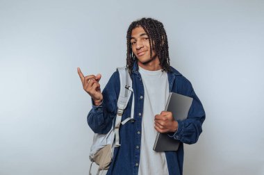 Saçları örgülü genç bir öğrenci elinde dizüstü bilgisayar ve sırt çantası ile güvenle ayakta duruyor. Önündeki okul maceralarına hazırlık ve heyecanı yansıtan neşeli bir jest yapıyor..