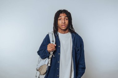 Genç bir adam sırt çantası taşıyan basit bir arkaplan karşısında duruyor. Kendine güveni tam, rahat bir duruşu ve günlük kıyafetleri var. Şehirde yaratıcı bir güne hazır..