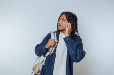 Genç bir adam telefonuyla konuşurken tuhaf bir ifadeyle ayakta duruyor. Sırtında bir sırt çantası taşıyor, sıradan bir kot ceket giyiyor, arka planda hafif bir dekoru var..
