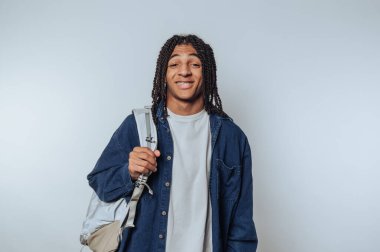 Neşeli genç bir adam sırtında bir sırt çantasıyla durur, dostça bir gülümsemeyle sevinçlerini ifade eder. Konfor ve tarz saçan basit bir kıyafet giyiyor..