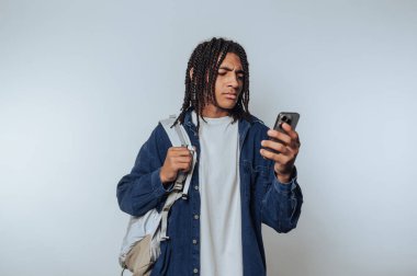 Genç bir adam akıllı telefonuna odaklanmış, gelişigüzel duruyor. Sırt çantası bir omuz üzerinde, şehir tarzını yansıtan rahat bir kıyafet giymiş. Arkaplan basit ve hafif.
