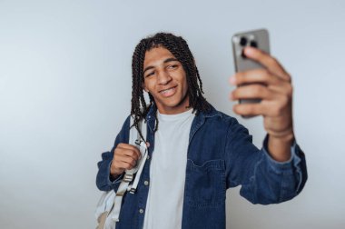 Genç bir adam, bir eliyle telefonunu, diğer eliyle sırt çantasını tutarak bir selfie çekerken parlak bir şekilde gülümsüyor. Gündelik tarzı, gençlik enerjisiyle dolu anın cazibesini arttırıyor..