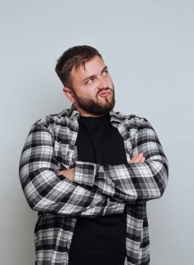 Sakallı bir adam kollarını kavuşturup dikkatlice yukarı bakar. Kareli fanilanın altına siyah bir gömlek giyer. Arkaplan hafif, sakin bir atmosfer yaratıyor..