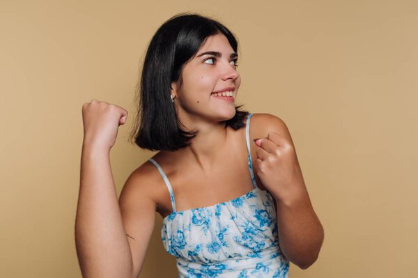 A young woman smiles brightly while making a playful fist pump with her arms raised. She radiates happiness and confidence against a warm, neutral background.
