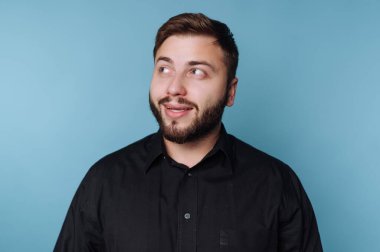 Sakallı genç bir adam parlak mavi bir arka plana karşı şakacı bir incelik sergiliyor. Şık bir siyah gömlek giyiyor, yaratıcı bir atmosferde kendine güven ve cazibe yayıyor..