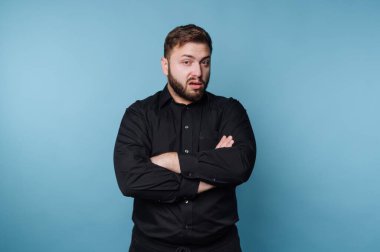 Parlak mavi bir ortamda, siyah gömlekli bir adam kollarını kavuşturarak güvenle ayakta duruyor. İfadesi düşünceli olduğunu gösteriyor. Davranışlarına derinlik katıyor..