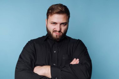 Kolları çapraz duran bir adam ciddi ama içsel bir ifade sergiliyor. Parlak mavi arkaplan, onun düşünceli tavırlarını vurgulayarak çarpıcı bir kontrast oluşturuyor..