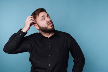 Sakallı bir adam siyah bir gömlek giyer ve kafasını kaşırken şaşkın bir yüz ifadesi gösterir. Yukarıya, derin düşüncelere, parlak mavi bir arkaplana bakıyor..