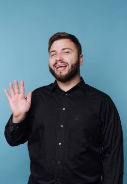 A cheerful man, wearing a black shirt, greets warmly with a wave and a big smile. The simple blue backdrop adds to the lively and friendly atmosphere of the moment.