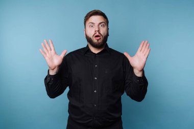 Bir adam canlı bir mavi zemine karşı duruyor, elleri havada ve ağzı açık şaşkınlık içinde. Siyah gömleği şaşkınlığını tamamlıyor, çarpıcı bir görüntü yaratıyor..