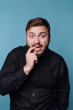 Sakallı bir adam tek parmağıyla ağzına dokunarak şaşkınlık ve merak gösterir. Parlak mavi bir arkaplanı olan siyah bir gömlek giyer, neşeli bir anı yakalar..