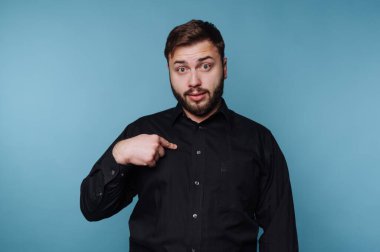 Genç bir adam göğsüne şaşkın bir ifadeyle işaret ederek parlak mavi bir arkaplan karşısında duruyor. Koyu renk gömleği, neşeli atmosfere zıtlık katıyor ve entrikaları cezbediyor..