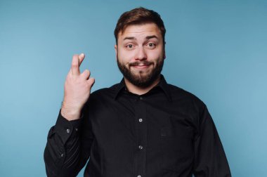 Bir adam, neşeli bir gülümseme ile parmaklarını çaprazlayarak, yumuşak mavi bir arkaplana karşı güvenle duruyor. Siyah gömleği parlak renkle çelişiyor, neşeli bir hava ve pozitif bir hava yayıyor..