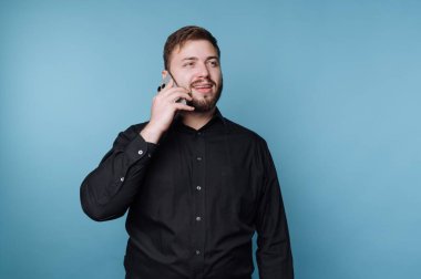 Siyah gömlekli gülümseyen bir adam telefonuyla konuşuyor. Canlı mavi arka plan neşeli atmosferi güçlendiriyor. Bu da olumlu bir konuşma olduğunu gösteriyor..