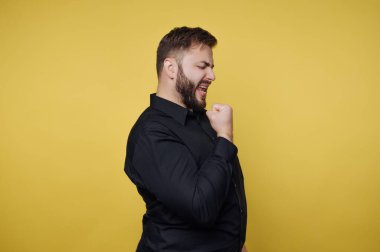 Siyah gömlek giyen sakallı bir adam, bu enerjik anda saf neşe ve heyecan gösteren canlı sarı bir arka plana karşı muzaffer bir yumrukla kutlama yapar..