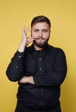 A man with a beard stands confidently in a black shirt, making a quirky gesture with his hand while smiling slightly. The vibrant yellow backdrop adds warmth and energy to the scene.
