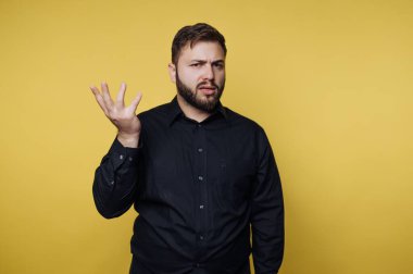 Siyah gömlekli, çarpıcı sarı bir arka plana karşı duran bir adam. Bir soru jestinde bir elini kaldırıp, anlık bir merak anını yakalarken ifadesi kafa karışıklığına yol açıyor..