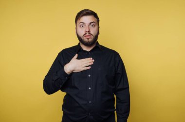 Bir adam ürkmüş bir ifadeyle ayakta duruyor, bir eli göğsünde, sürpriz bir şekilde. Koyu renk gömleği parlak sarı arka planla cesurca zıtlaşıyor, dramatik sahneyi güçlendiriyor..