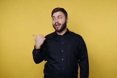 A cheerful man stands against a vibrant yellow wall, wearing a black button-up shirt and playfully giving a thumbs up while making a funny expression, radiating enthusiasm and positivity.