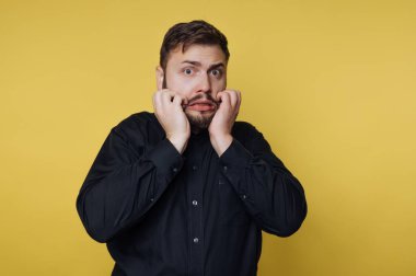 Siyah gömlekli genç bir adam abartılı bir şok ve şaşkınlık bakışı sergiliyor. Yüzündeki geniş gözleri ve elleri, koyu sarı bir zemin üzerine kurulu komik bir reaksiyonu işaret ediyor..