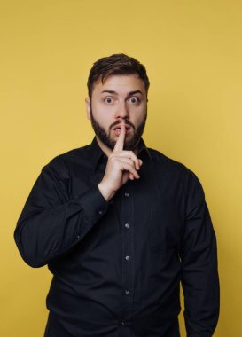 Siyah düğmeli gömlekli, sürpriz bir adam, parlak sarı bir duvara karşı duruyor, bir gizlilik jesti olarak parmaklarını dudaklarına koyuyor. Geniş gözleri merak ve haylazlık taşıyor..