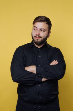 A man stands with crossed arms, showcasing a confident demeanor. The bright yellow backdrop contrasts with his black shirt, adding a striking visual element to the scene.
