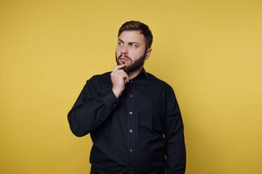 Siyah gömlekli, parlak sarı bir duvara karşı düşünceli bir adam duruyor. Düşüncelere dalmış, bir eli çenesinde, merak ve yansıma havası yayıyor..