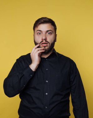 A man stands in a studio, eyes wide and mouth agape, showcasing a surprised expression. His hand rests on his face, reflecting astonishment in a bright yellow setting.