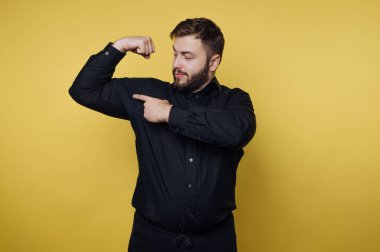 Siyah bir gömlek giymiş, kaslarını kasan bir adam, parlak sarı bir arka planda duruyor. İfadesi, spor için gurur ve coşku saçıyor..