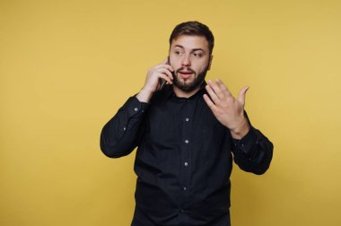 Siyah gömlekli bir adam, parlak sarı bir duvara karşı duruyor, telefonda tutkuyla tartışıyor ve el hareketleriyle noktalarını vurguluyor..