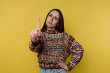 Genç bir kadın, sanki bekleyecekmiş gibi parmağını göstererek neşeli bir yüz ifadesi sergileyen, parlak sarı bir duvara karşı güvenle duruyor. Onun desenli süveteri anı güzelleştiriyor..