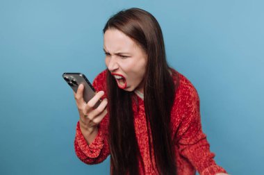 A young woman stands with long hair and an intense expression, yelling into her smartphone. She wears a bright red sweater, showing her strong emotions. The colorful backdrop adds to the moment.