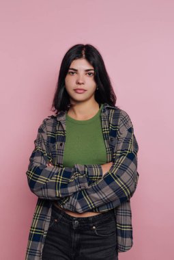 Yeşil bluz ve ekose gömlek giyen kendine güvenen genç bir kadın çapraz kollarla ayakta duruyor. İfadesi güç ve bireysellik yayıyor. Eşsiz tarzını pembe bir arka plana vurguluyor..