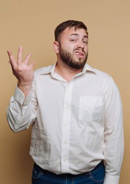 Beyaz gömlekli bir adam bej bir duvara karşı duruyor ve bir belirsizlik jesti ile elini kaldırıyor. Yüzündeki ifade kafa karışıklığını gösteriyor..