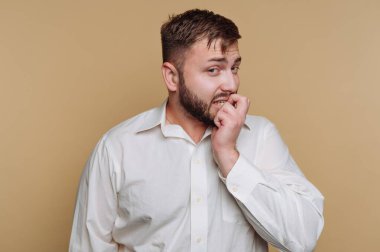 A man in a white shirt stands against a warm yellow backdrop, anxiously biting his nails. His expression reflects concern or apprehension, creating a relatable moment of vulnerability.