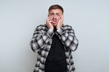 In a contemporary room, a young man stands with wide eyes and clenched hands on his face, showcasing intense emotion. His plaid shirt contrasts with the plain backdrop, capturing a moment of surprise.