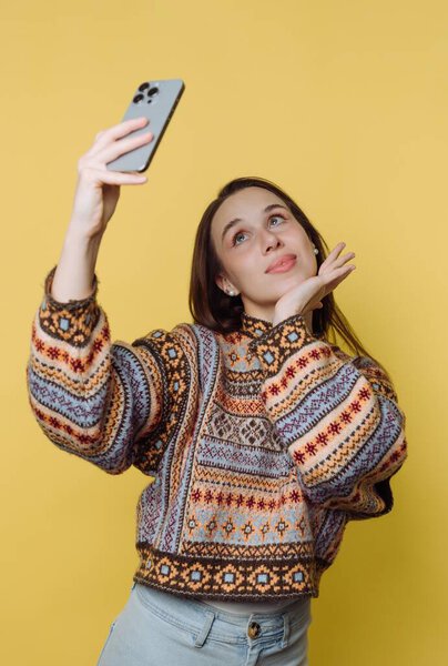 A young woman takes a fun selfie while playfully sticking her tongue out. She wears a colorful, patterned sweater, adding to the cheerful vibe of the bright yellow backdrop.