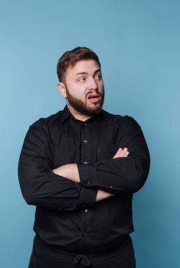 Genç bir adam kollarını kavuşturup düşünceli bir ifade sergileyerek özgüvenle ayakta duruyor. Siyah bir gömlek giyiyor, parlak mavi bir arkaplanı var, koyu bir kontrast oluşturuyor..