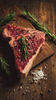 Top view flat lay of raw T-bone steak with rosemary, salt, and pepper on rustic wooden background with copy space, warm natural light, realistic texture, gourmet cooking concept, professional style for website or print use.