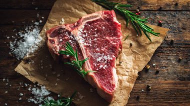 Top view flat lay of raw T-bone steak with rosemary, salt, and pepper on rustic wooden background with copy space, warm natural light, realistic texture, gourmet cooking concept, professional style for website or print use.