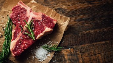 Top view flat lay of raw T-bone steak with rosemary, salt, and pepper on rustic wooden background with copy space, warm natural light, realistic texture, gourmet cooking concept, professional style for website or print use.
