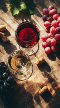 Top view flat lay of red and white wine glasses on wooden table, scattered grapes and corks, blurred background with copy space, warm natural light, elegant beverage concept, realistic style for website or print use.