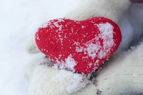 Mãos em mitenes de malha brancas com um coração vermelho em um dia de inverno em um contexto de neve. — Fotografia de Stock