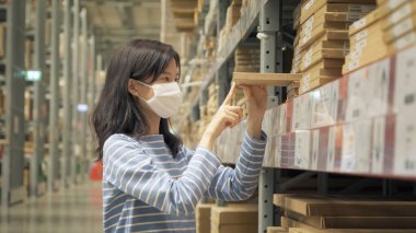 Büyük bir depo perakende mağazasında maske takan Asyalı kadın çalışıyor ya da raflarda alışveriş yapıyor. Mobilya ve ev aksesuarları. Ekommerce ve lojistik kargo. Yaşam biçimi