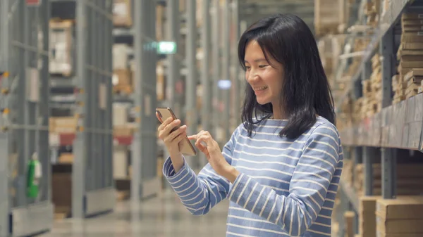 Asian woman using a smart phone or shopping on shelves at large warehouse retail store industry. Rack of furniture and home accessories. Interior of cargo in ecommerce and logistic concept. Technology