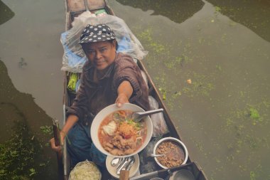 Marketteki kadın Damnoen Saduak Yüzen Market 'te ya da Amphawa' da erişte veriyor. Yerel halk, Tayland 'ın Ratchaburi bölgesindeki teknelerde meyve ve geleneksel yiyecekler satıyor. Meşhur Asya turistik cazibesi.