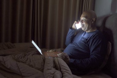 Mutlu gülümseme işleri, siyahi adam, Afro-Amerikalı insanlar evden çalışıyor ve telefonda iş arkadaşlarıyla konuşuyor, karantinada video konferansı gece yarısı yatak odasında yatak odasında. Yaşam biçimi