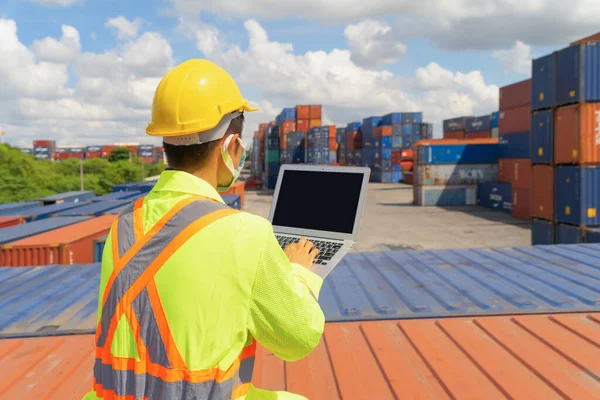 Asian logistic worker engineer man using a laptop computer, working in cargo container warehouse industry factory site in export, import, and transportation concept. Business people. Technology device