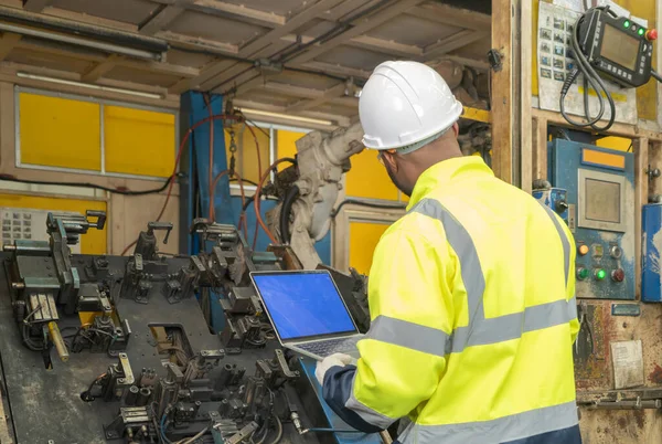 Black African American man, an engineer or worker using a laptop notebook computer device to control smart robot welding hand system automate manufacturing machine engine in factory, industry. People.